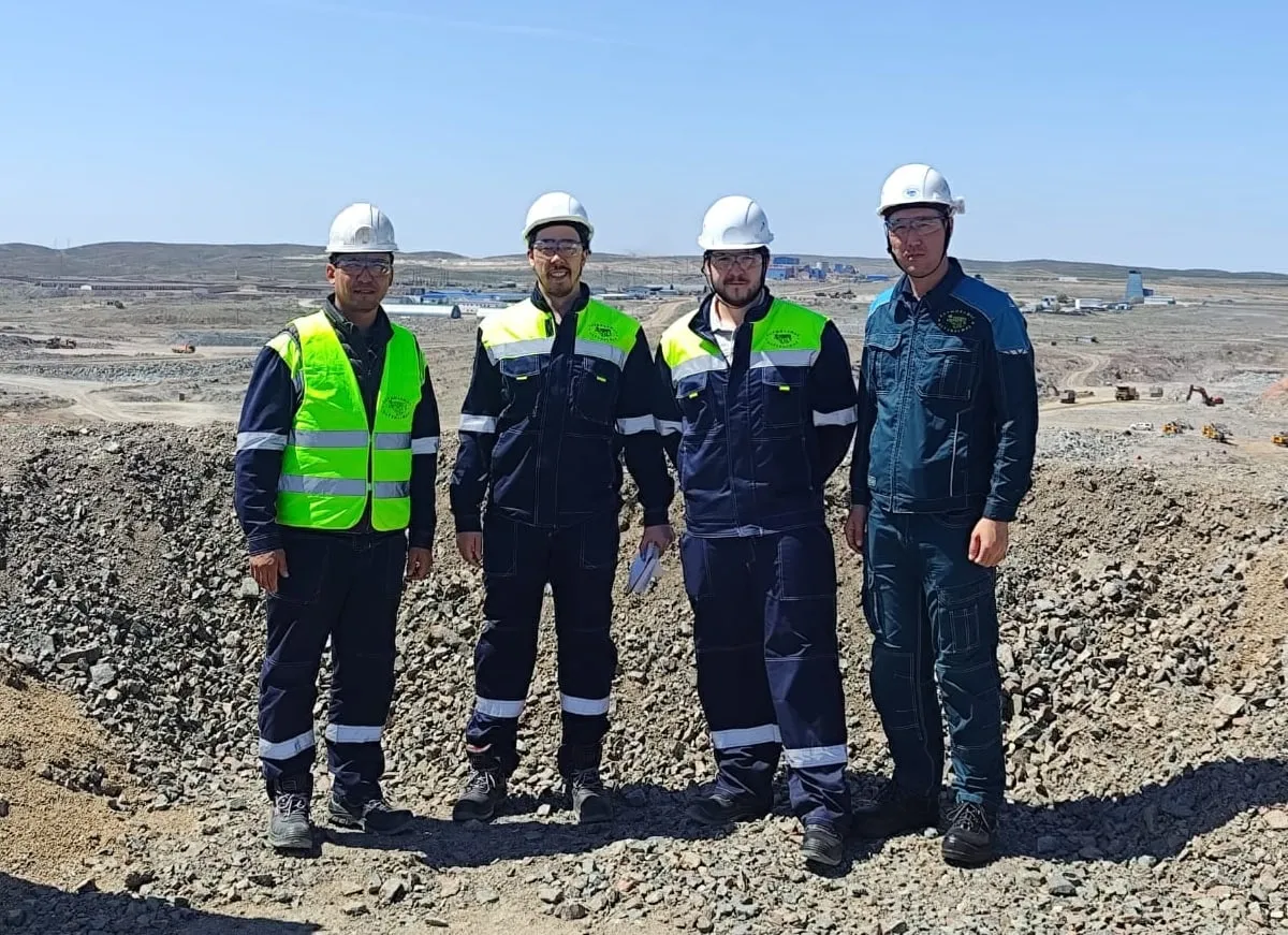 Minebright engineers in hard hats, safety glasses and hi-vis workwear on broken rock above an open-pit mine, haul roads and plant buildings behind them.